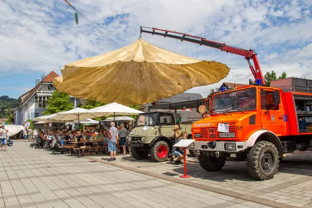 Unter dem großen beigen Lastenfallschirm sitzen die Gäste an den Bierbänken; davor ein oranger Feuerwehr-Unimog mit Ladekran und ein grüner Unimog.