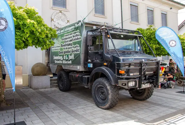 Dunkler Unimog mit grünem Einladungsbanner zum Treffen steht vor dem historischen Biberacher Rathaus, flankiert von zwei blauen Unimog-Club-Fahnen.