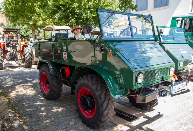 Ein grüner Oldtimer-Unimog mit auffällig roten Felgen steht offen auf dem Kopfsteinpflaster, ein Kind sitzt am Steuer.