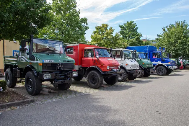 Mehrere Unimog in Grün, Rot, Weiß und Blau stehen aufgereiht auf dem Festplatz und zeigen die Vielfalt der Mercedes-Geländefahrzeuge.